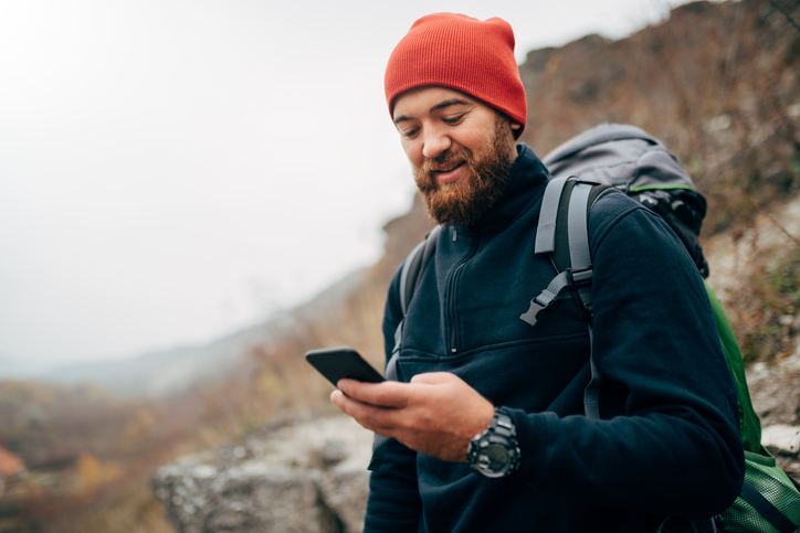 Young bearded man smiling and sending messages for his family from his cellphone, during hiking in mountains. Traveler bearded man in red hat using mobile phone application